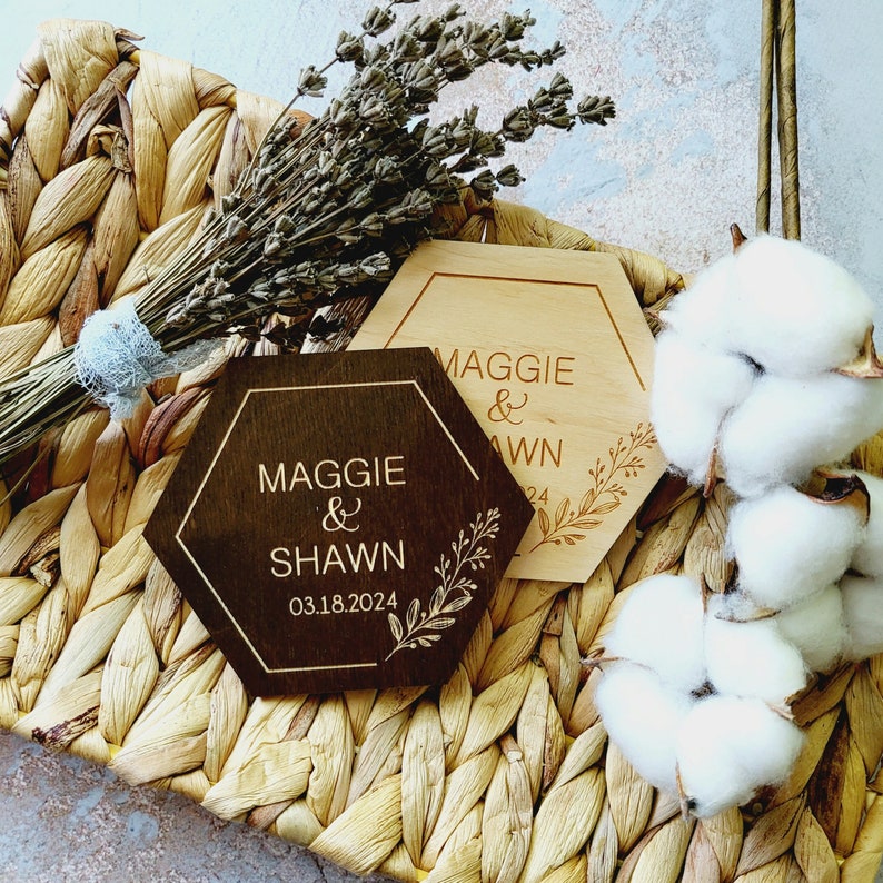 May include: Two hexagonal wooden signs with the text "Maggie & Shawn" and the date "03.18.2024" on one sign and "Maggie & Shawn" on the other. The signs are on a woven wicker surface with dried lavender and cotton balls.