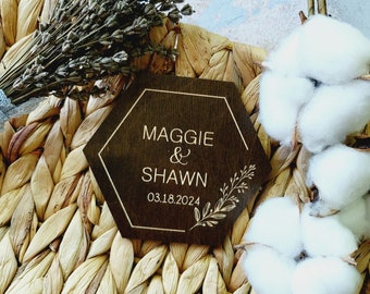 May include: A hexagonal wooden sign with a gold border and the text "Maggie & Shawn 03.18.2024" in gold lettering. The sign is on a woven straw background with white cotton balls.