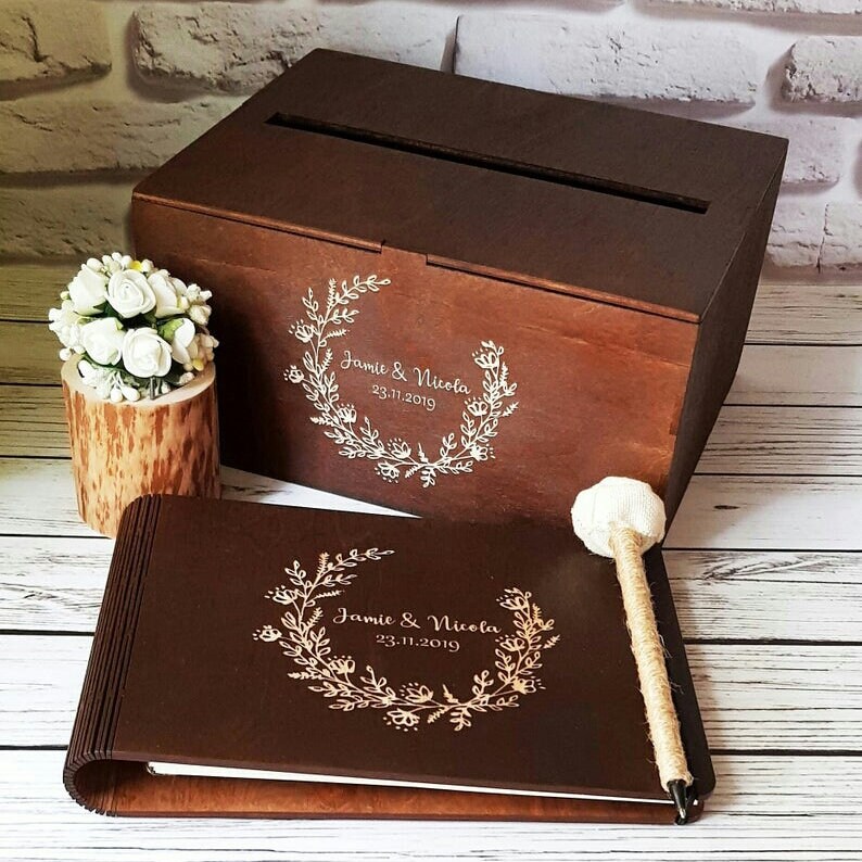 Op de afbeelding: Een houten gastenboek en kaartenbox voor een bruiloft met een bloemmotief en de namen Jamie & Nicola en de datum 23.11.2019 gegraveerd op de voorkant.