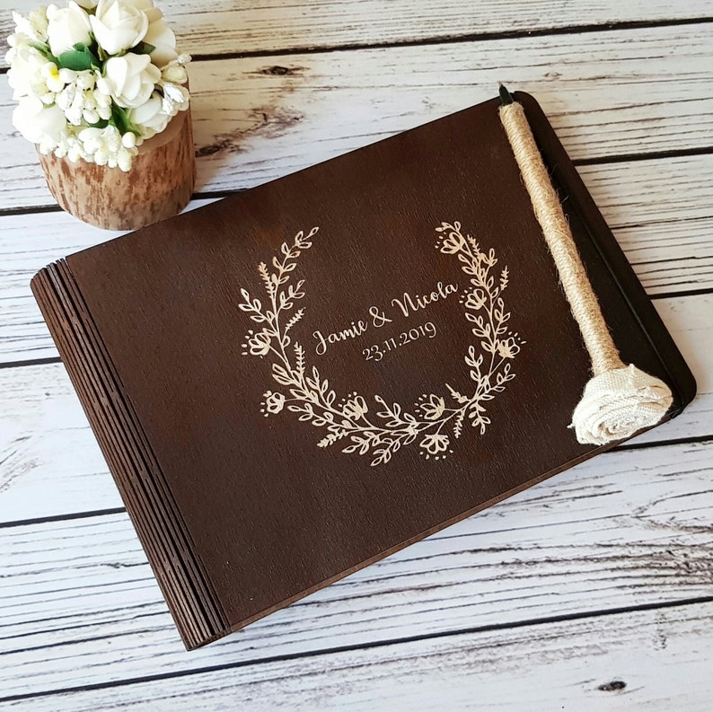 Op de afbeelding: Een donkerbruin houten gastenboek met een bloemenkransontwerp en de tekst "Jamie & Nicola 23.11.2019" gegraveerd op de voorkant. Het boek is gesloten en heeft een rustieke touw sluiting met een witte stoffen bloem.