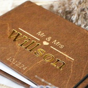 Op de afbeelding: Een houten gastenboek met een gouden inscriptie die "Mr. & Mrs. Wilson" en de datum "10.12.24" luidt.