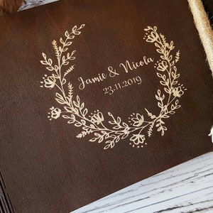 Op de afbeelding: Een donkerbruin houten fotoalbum met een ontwerp van een bloemenkrans en de tekst "Jamie & Nicola 23.11.2019" gegraveerd in goud.
