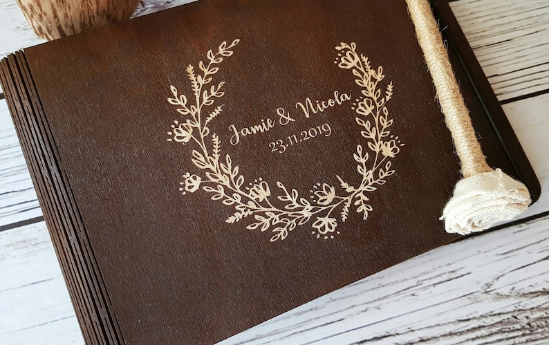 Op de afbeelding: Een donkerbruin houten fotoalbum met een ontwerp van een bloemenkrans en de tekst "Jamie & Nicola 23.11.2019" gegraveerd in goud.