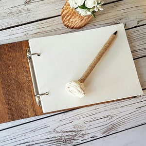Op de afbeelding: Een houten gastenboek met een witte papieren inleg en een ringband. Een pen met een stoffen roos aan het uiteinde ligt op het open boek.