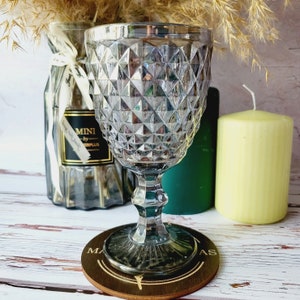 May include: A clear glass goblet with a diamond pattern sits on a wooden coaster with the word "MAS" printed on it. The goblet is on a white wooden surface with two candles in the background.