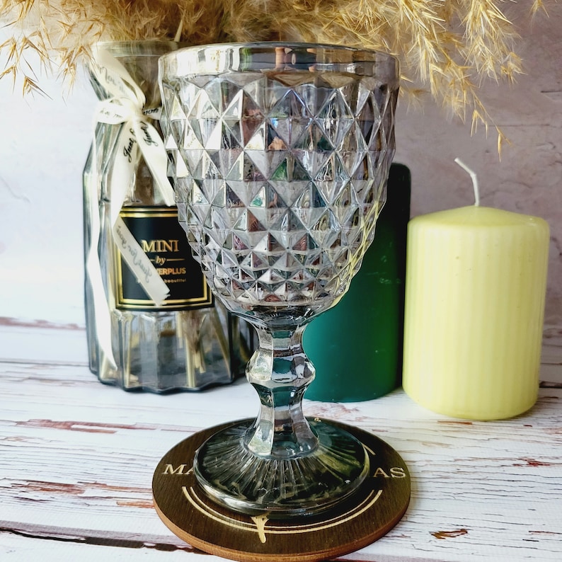 May include: A clear glass goblet with a diamond pattern sits on a wooden coaster with the word "MAS" printed on it. The goblet is on a white wooden surface with two candles in the background.