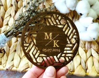 May include: A round wooden coaster with a geometric design cut out. The center of the coaster features a circle with the initials "M/K" and the date "05.07.2024" engraved in a lighter wood.