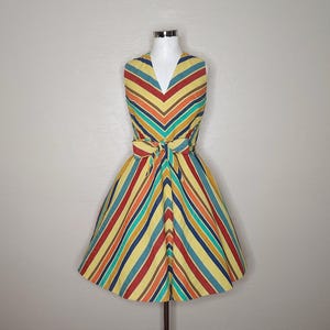 May include: A sleeveless, knee-length dress with a V-neck and a fitted waist. The dress features a colorful striped pattern in yellow, red, blue, orange, and brown. A matching fabric belt is tied at the waist. The dress is displayed on a mannequin.