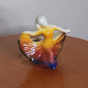May include: A ceramic figurine of a dancer with a yellow, orange, red, and blue dress. The figurine is in a dancing pose with arms outstretched and a skirt flowing behind her.