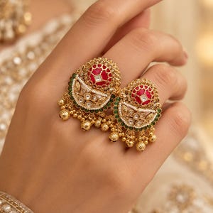 May include: A gold-toned ring featuring two ornate, semi-circular motifs. Each motif contains a central red circular inlay, surrounded by white stones, a border of small green beads, and dangling gold-toned spheres along the bottom edge.