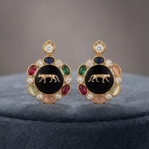 May include: A pair of gold-toned earrings featuring a central black circular inlay with a gold panther silhouette. The center is surrounded by a border of alternating oval-shaped multicolored gemstones and clear round stones, topped with a single diamond.