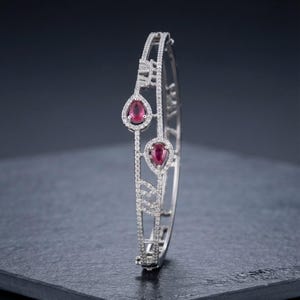 Peut inclure: Un bracelet jonc articulé de couleur argentée orné de deux pierres rouges en forme de poire, entourées de petites pierres claires. La structure métallique ajourée présente un motif géométrique entièrement serti de minuscules pierres claires.