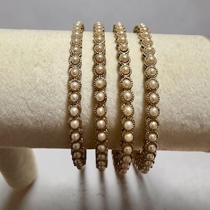 May include: Four gold-toned metal bangles, each featuring a continuous row of small, round, cream-colored faux pearls set within a textured, scalloped metal frame. The bracelets are arranged vertically, showcasing the uniform bead design.