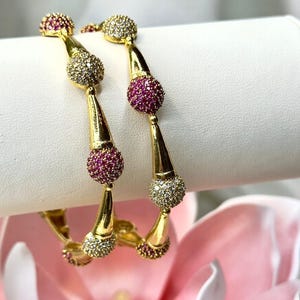 May include: Two gold bangles with alternating ruby and clear crystal studded spheres. The bangles have a unique design with a teardrop shape and are displayed on a white surface with a pink flower in the background.