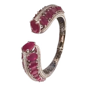 May include: A silver-toned open cuff bracelet featuring a row of oval-cut deep red rubies set along the center. The edges of the bracelet are lined with small, clear pavé-set stones, and the inner band displays a detailed, intricate lattice metalwork pattern.
