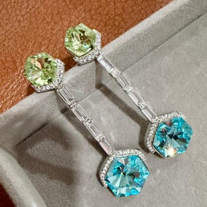 May include: A pair of elegant earrings featuring a light green and a blue hexagonal gemstone, each surrounded by a halo of small, clear stones. The earrings have a long, slender silver bar with embedded clear stones.