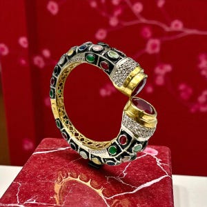 May include: A gold and silver bracelet with a black enamel band, adorned with colorful gemstones and diamonds. The bracelet features two large, oval-cut red gemstones at the ends. The jewelry is displayed on a red marble surface.