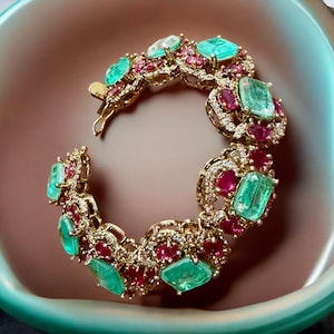 May include: A gold bracelet featuring emerald and ruby gemstones. The bracelet has a repeating pattern of large, rectangular emeralds and smaller, round rubies, set in a gold filigree design. The bracelet is displayed in a light pink bowl.