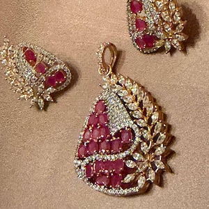May include: A jewelry set consisting of a pendant and two matching earrings. Each piece features a gold-tone metal base adorned with clusters of oval-shaped deep red stones and numerous small, clear, marquise-cut stones arranged in a leaf pattern.