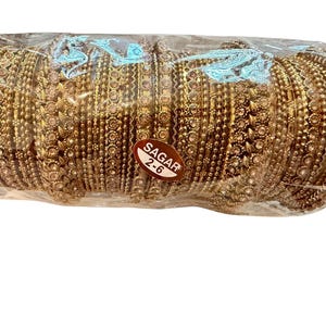 May include: A bundle of gold-colored bangles, intricately designed with floral and beaded patterns. The bangles are wrapped in clear plastic. A small, oval sticker with the text "SAGAR 2-6" is visible.