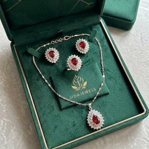 May include: A jewelry set featuring a necklace, a ring, and a pair of earrings, each with a pear-shaped red stone surrounded by a halo of small white stones. The set is presented in a dark green velvet box with the text JIDAJEWELS printed in gold.
