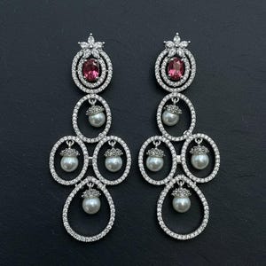 May include: Elegant chandelier earrings with a floral top, a pink gemstone, and dangling pearls. The earrings are silver-toned and encrusted with small, sparkling crystals. The design is symmetrical and eye-catching.