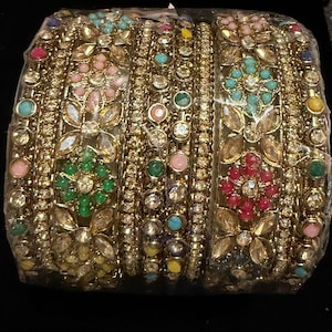 May include: A set of gold-toned bangles encased in clear plastic packaging. The bracelets feature intricate metalwork, clear rhinestones, and small round beads in various colors including pink, green, yellow, and blue arranged in floral patterns.