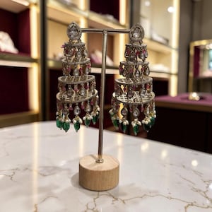 May include: A pair of tiered chandelier earrings featuring a metallic lattice structure adorned with clear teardrop stones, small pink beads, and dangling green teardrop-shaped accents. The earrings are displayed on a silver-toned metal stand.