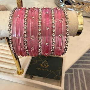 May include: A set of translucent pink bangles arranged on a display stand. The collection includes several smooth pink bracelets interspersed with silver-toned metal bangles encrusted with clear rhinestones. A small black card displays the text JIDAJEWELS.