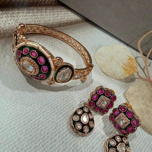 May include: A gold-toned hinged bangle bracelet and a pair of matching drop earrings. The jewelry features dark green enamel, vibrant pink oval stones, and clear central stones, all accented with small white beads along the bangle edge.