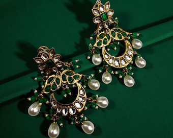 May include: A pair of gold-toned earrings with green and white accents. The earrings feature a floral design at the top, green leaf-shaped details, and dangling white pearl-shaped beads. The earrings are set against a dark green background.