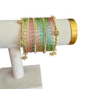 May include: A collection of colorful bangles in shades of green, pink, and yellow, displayed on a white jewelry stand. The bangles are adorned with gold accents and small dangling charms. A central bangle is embellished with clear crystals.