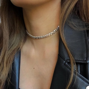 May include: A silver chain choker necklace. The necklace is a curb chain style, with a series of interlocking links. The necklace is worn around the neck. The person is wearing a black top and a black leather jacket.