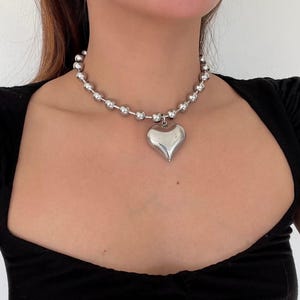 May include: A silver chain necklace with a large, heart-shaped pendant. The chain is made of small, round beads.
