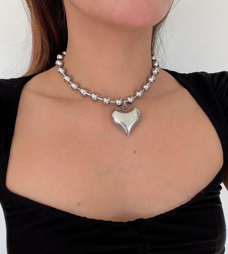 May include: A silver chain necklace with a large, heart-shaped pendant. The chain is made of small, round beads.