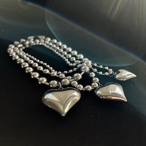 May include: A silver chain necklace with three heart-shaped pendants. The chain is made of small, round beads.