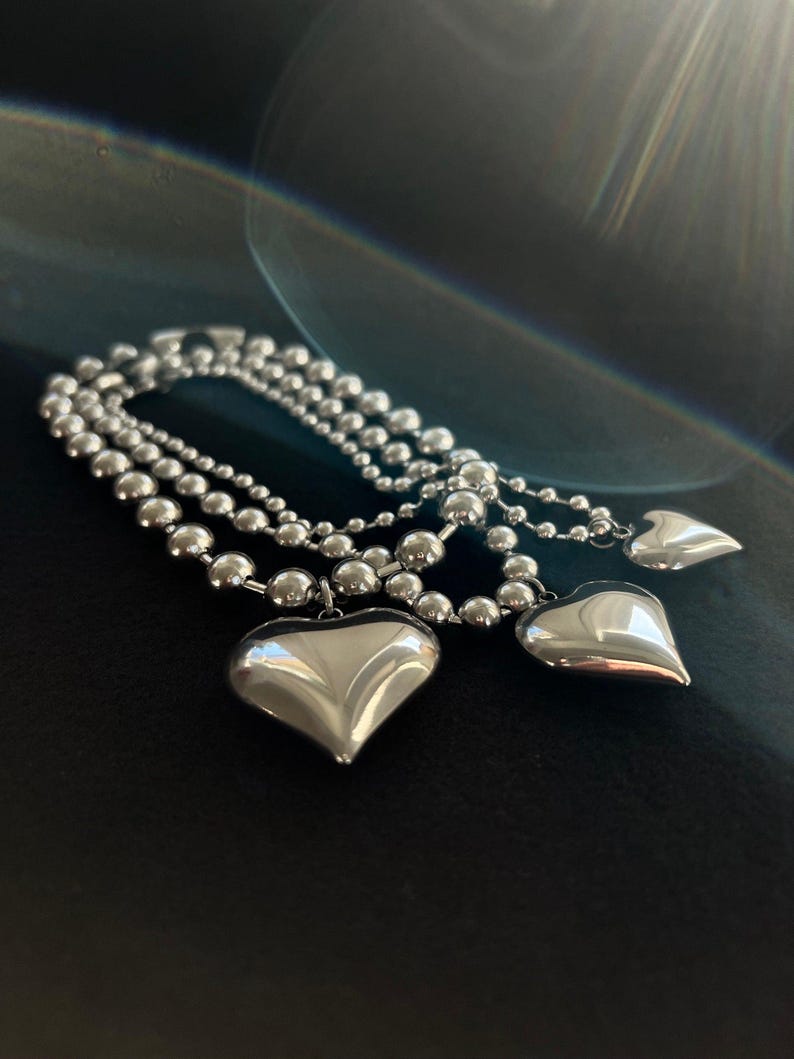 May include: A silver chain necklace with three heart-shaped pendants. The chain is made of small, round beads.