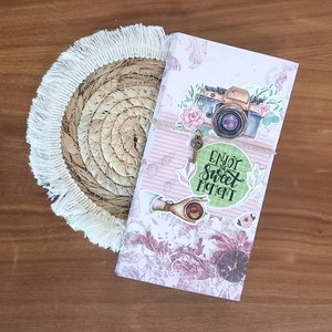 May include: A pink and white journal with a floral design and a camera sticker. The journal has a key charm attached to it and a green circle sticker that says "Enjoy the Sweet Moment".
