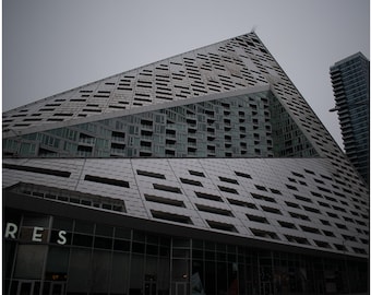 May include: A modern, geometric building with a triangular design and a facade of gray panels and windows. The building's unique shape and architecture stand out against a cloudy sky. The word "RES" is visible on the building.