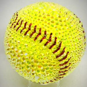May include: A bedazzled softball, primarily yellow with red stitching. The softball is covered in small, sparkling crystals, giving it a shimmering appearance. The red stitching is visible, and the ball is displayed on a clear stand.