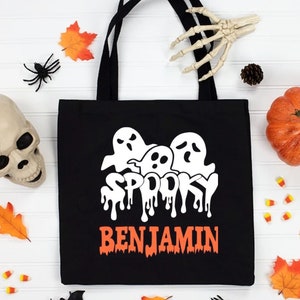 Peut inclure: Un sac fourre-tout noir avec un motif blanc représentant trois fantômes au-dessus du mot SPOOKY dans une police dégoulinante, avec le nom BENJAMIN imprimé en orange en dessous. Le design est entouré d'un crâne, d'une citrouille et de feuilles.