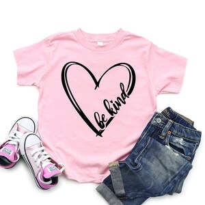 May include: Pink t-shirt with a black heart and the text "be kind" inside the heart. The shirt is paired with blue jeans and pink sneakers.