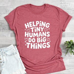 May include: A red t-shirt with the text "HELPING TINY HUMANS DO BIG THINGS" in white. The shirt is paired with denim shorts. A small green plant is placed to the side. The shirt is made of a soft material.