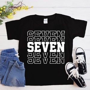 May include: Black t-shirt with white text that says "SEVEN" repeated in a stacked pattern. The text is outlined in black.