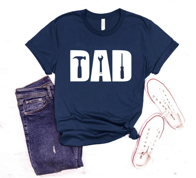May include: A navy blue t-shirt with the word "DAD" in white letters. The letters are decorated with a hammer, wrench, and screwdriver.