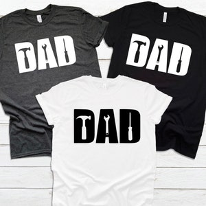 May include: Three t-shirts with the word "DAD" in white letters with a hammer, wrench, and screwdriver graphic inside each letter. The shirts are in grey, black, and white.