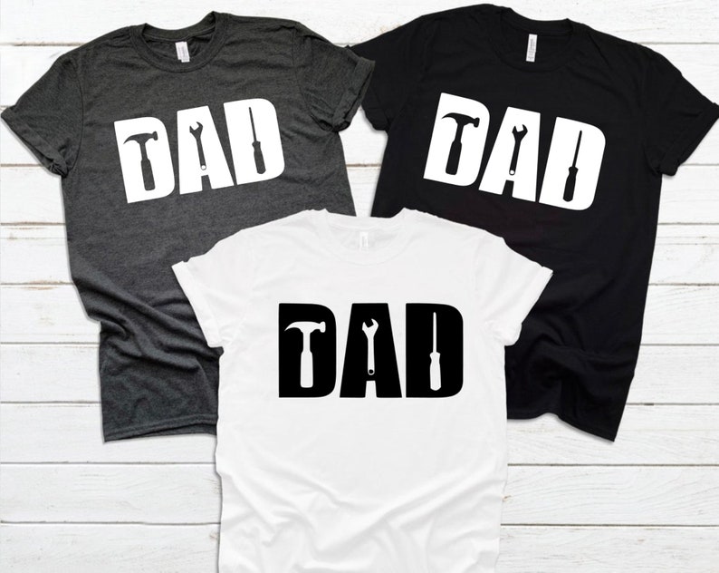 May include: Three t-shirts with the word "DAD" in white letters with a hammer, wrench, and screwdriver graphic inside each letter. The shirts are in grey, black, and white.