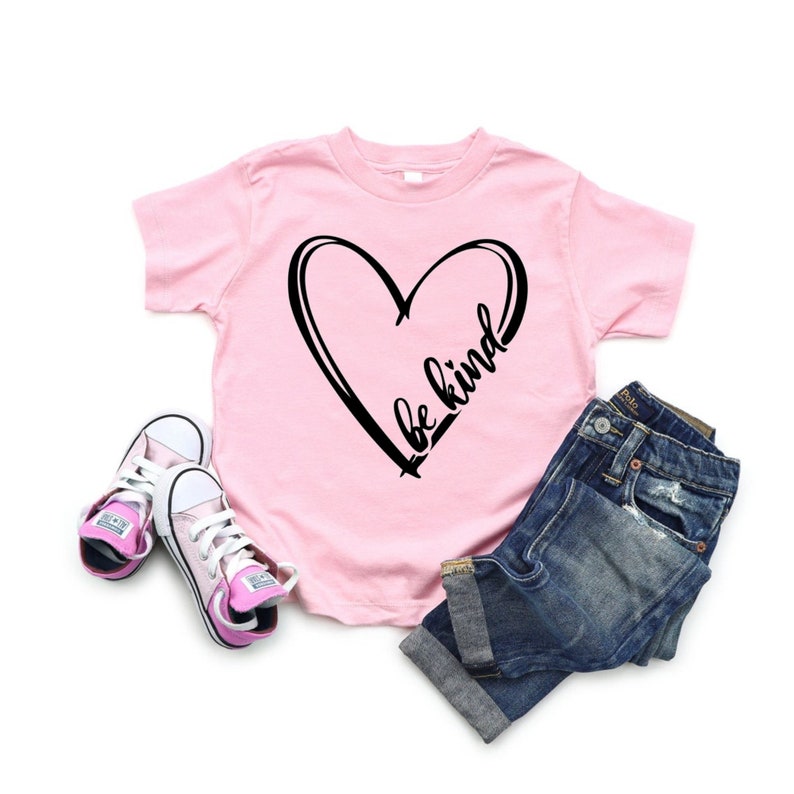 May include: Pink t-shirt with a black heart and the text "be kind" inside the heart. The shirt is paired with blue jeans and pink sneakers.