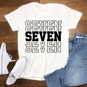 May include: White t-shirt with the word "SEVEN" printed in black letters, repeated five times in a stacked design. The shirt is laid flat on a wooden surface with a pair of white sneakers and blue jeans.