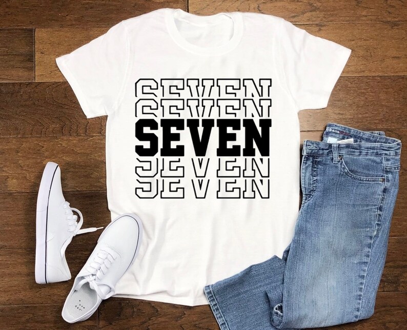 May include: White t-shirt with the word "SEVEN" printed in black letters, repeated five times in a stacked design. The shirt is laid flat on a wooden surface with a pair of white sneakers and blue jeans.